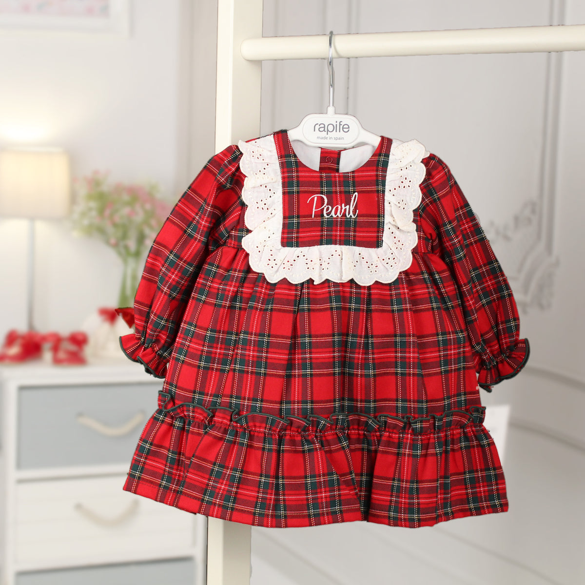 A baby girl's long-sleeve plaid dress in red, green, and black tartan, featuring a white scalloped eyelet collar with the name 'Pearl' embroidered in white. The dress is shown hanging on a white hanger labeled 'rapife' against a softly blurred nursery background.