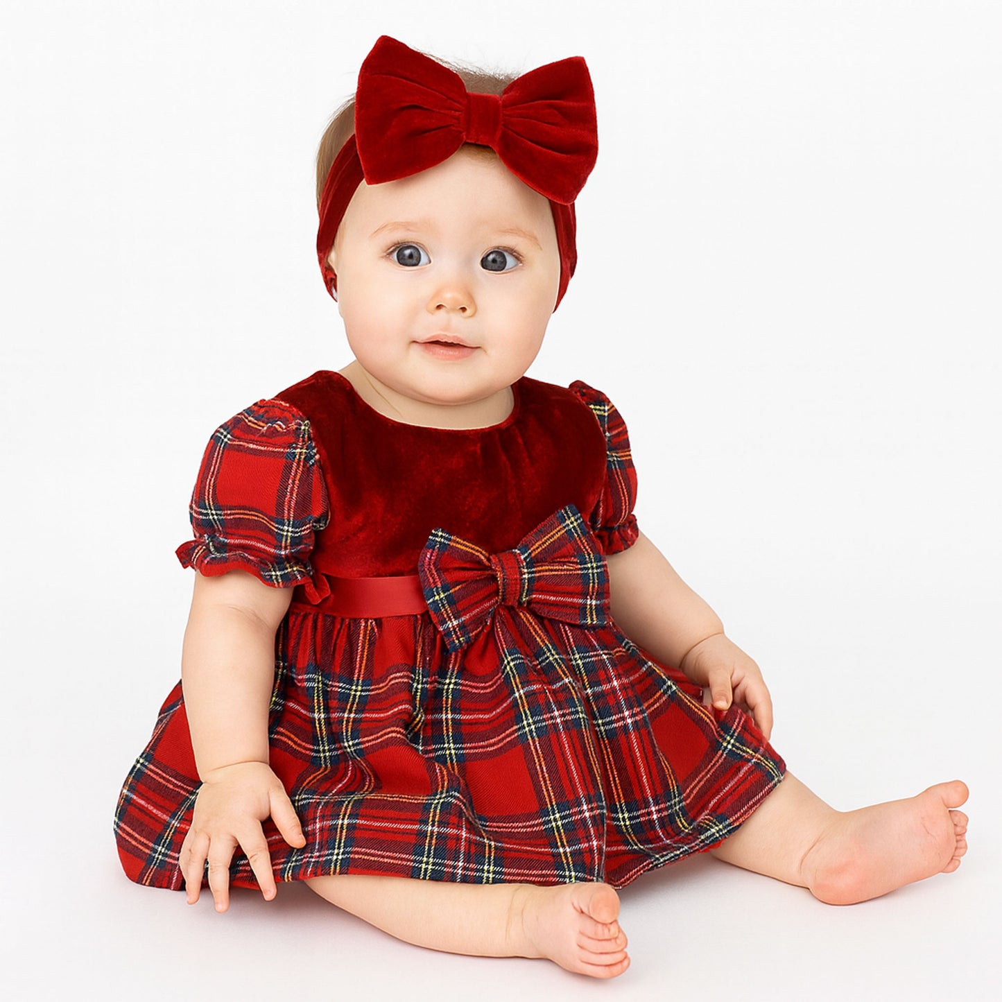 Baby wearing a red plaid dress with a large bow and matching headband on a white background