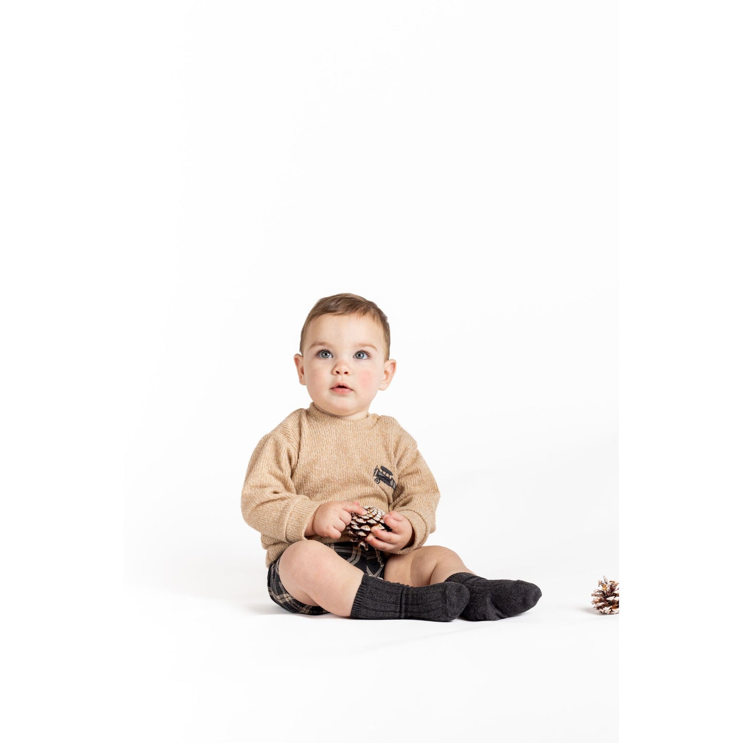 Baby sitting on a white background wearing a beige sweater and dark socks.