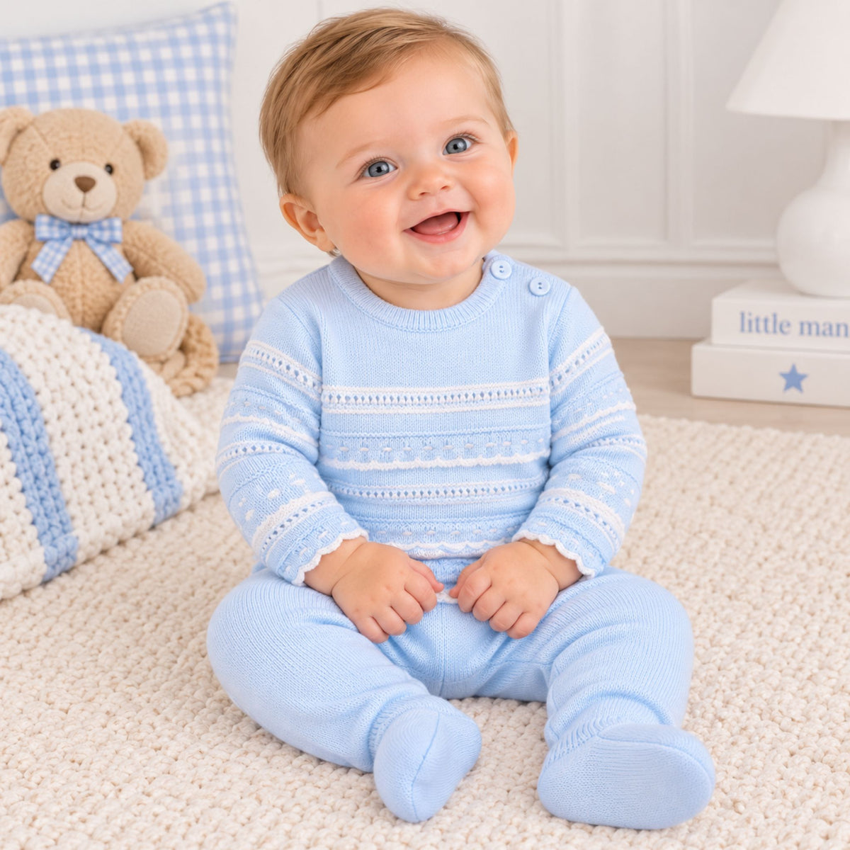 A baby sitting on a cream carpet wearing a light blue knit footed sleeper with white patterned stripes. The background features a teddy bear, a blue and white checkered pillow, and a white lamp.