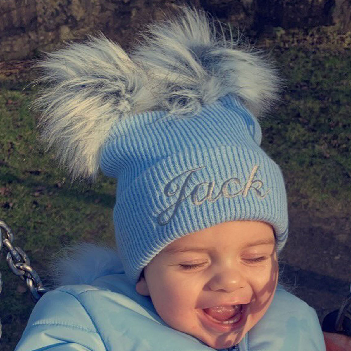 Child wearing a blue knit hat with a fluffy pom-pom, sitting outdoors.
