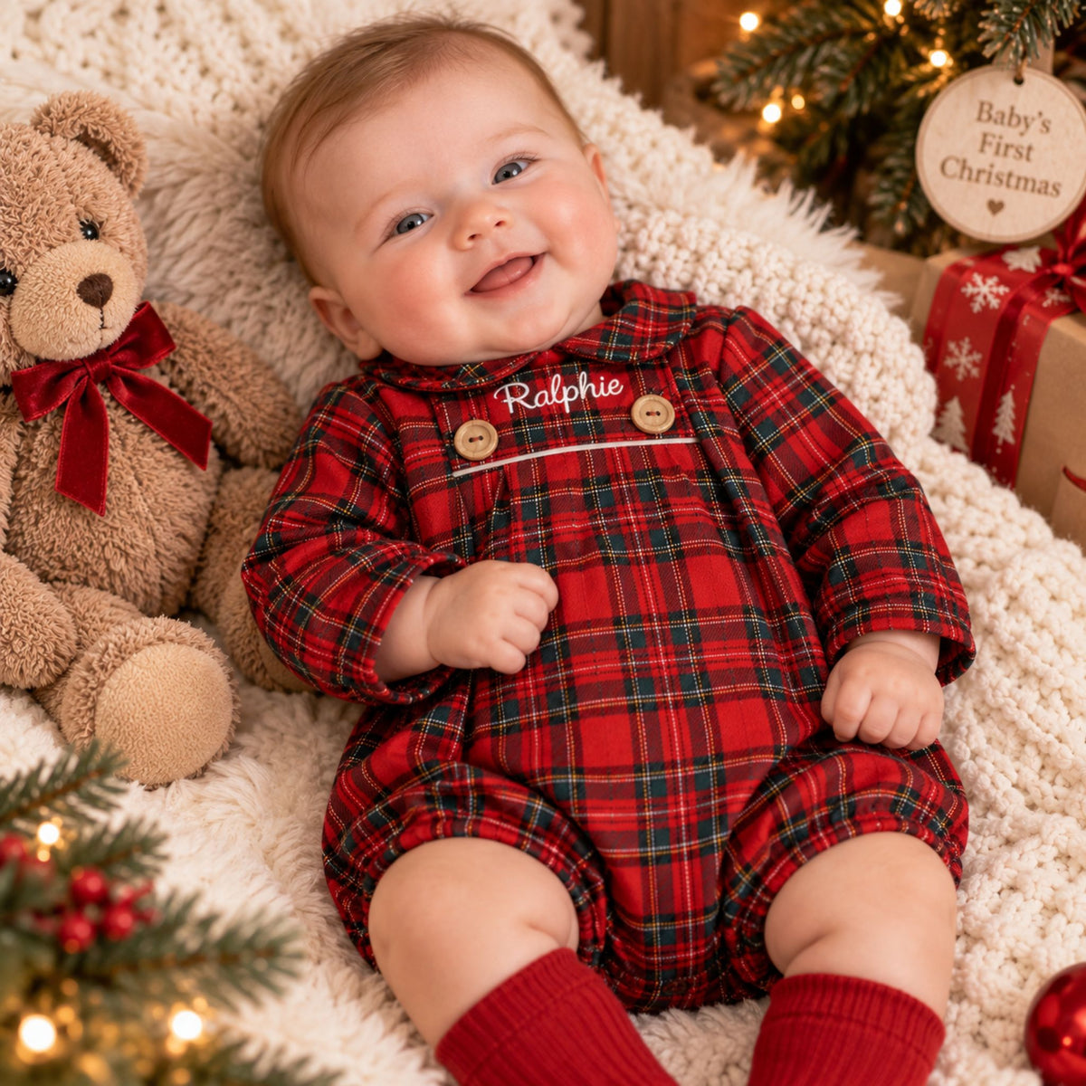 A baby lying on a cream knit blanket with a teddy bear, Christmas tree lights, and wrapped gifts in the background. The baby is wearing a red and green plaid long-sleeve romper with 'Ralphie' embroidered on the chest, wooden buttons, and red knit socks.