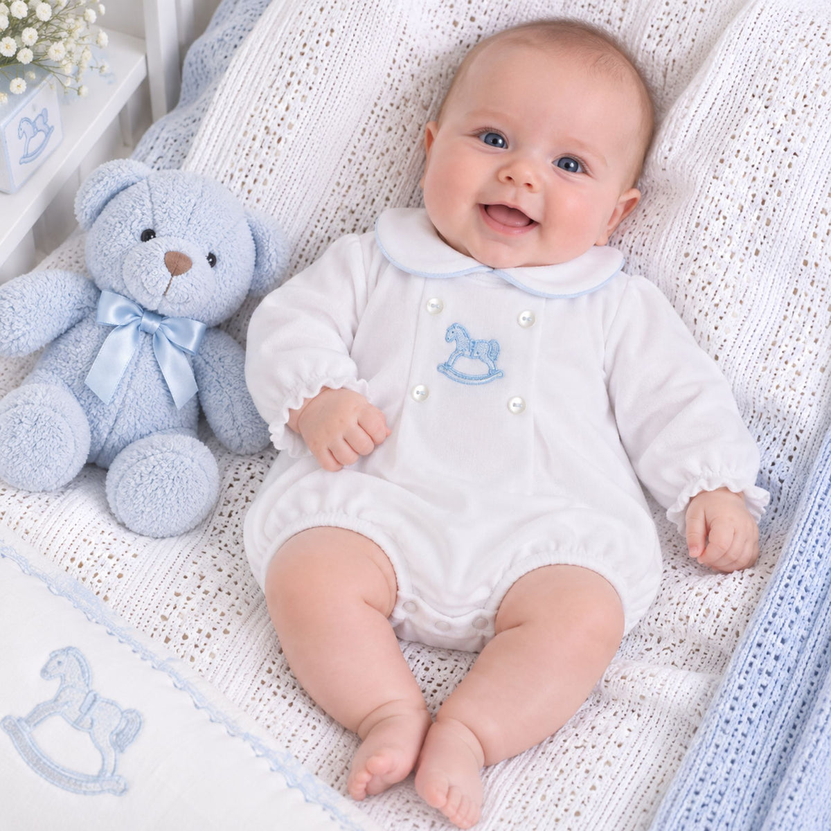 A smiling baby wearing a white long-sleeve romper with a light blue rocking horse motif and pearl-like buttons, lying on a white knit blanket next to a light blue teddy bear.