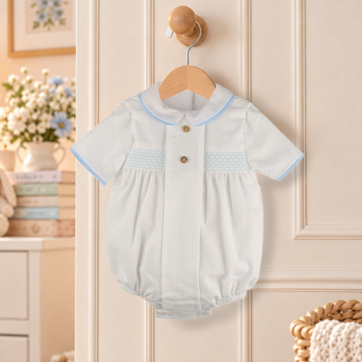 A baby short-sleeve romper in white with light blue trim and smocked chest detailing, hanging on a wooden hanger on a cream paneled door.