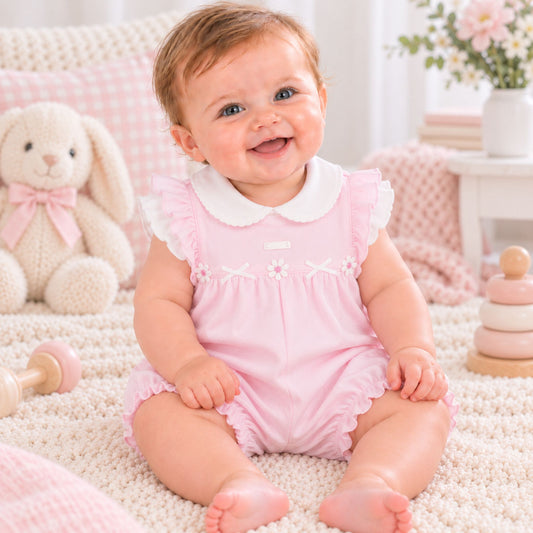 A smiling baby sitting on a cream textured rug wearing a light pink romper with a white Peter Pan collar. The background features a cozy nursery setting with a pink gingham pillow, a plush white bunny toy, and soft pastel decor.