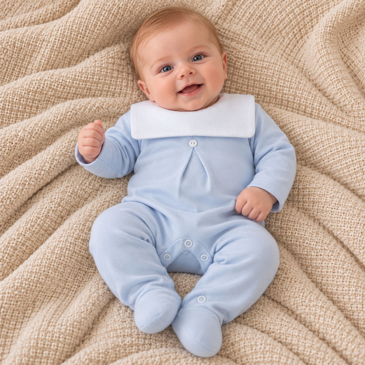 A baby lying on a textured beige blanket, wearing a light blue footed romper with a contrasting white collar.