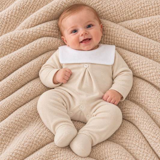 A baby lying on a textured beige blanket, wearing a cream-colored footed sleeper with a contrasting white collar.
