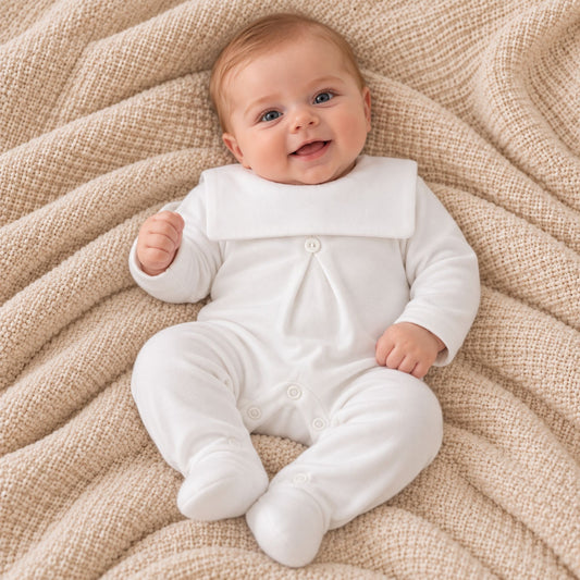 A baby lying on a textured beige blanket, wearing a white long-sleeve footed sleeper with a wide collar and snap closures.
