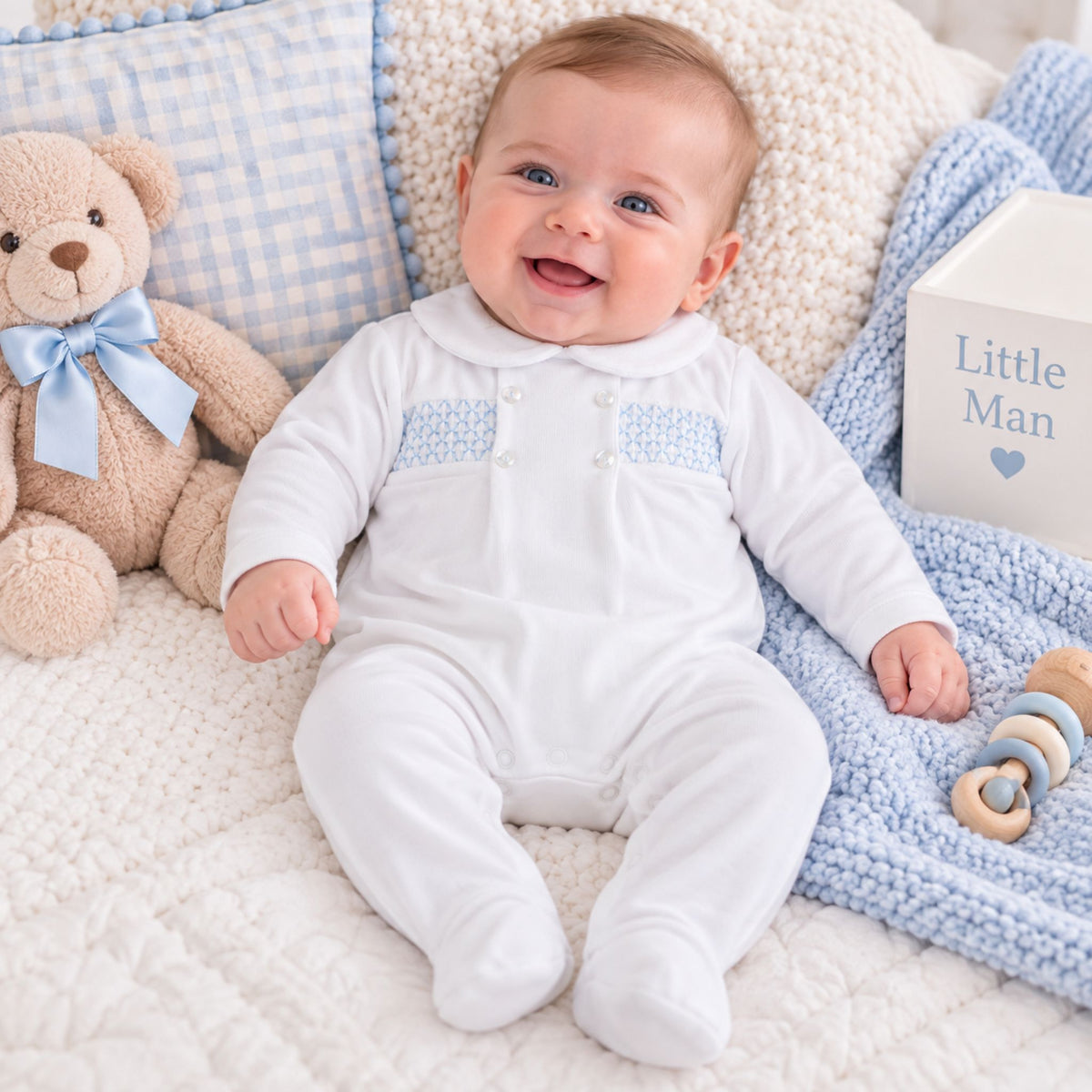 A smiling baby wearing a white long-sleeve footed romper with light blue knit chest panels and decorative buttons, lying on a white quilted blanket with a teddy bear, blue gingham pillow, blue knit blanket, and a wooden block that says 'Little Man'.