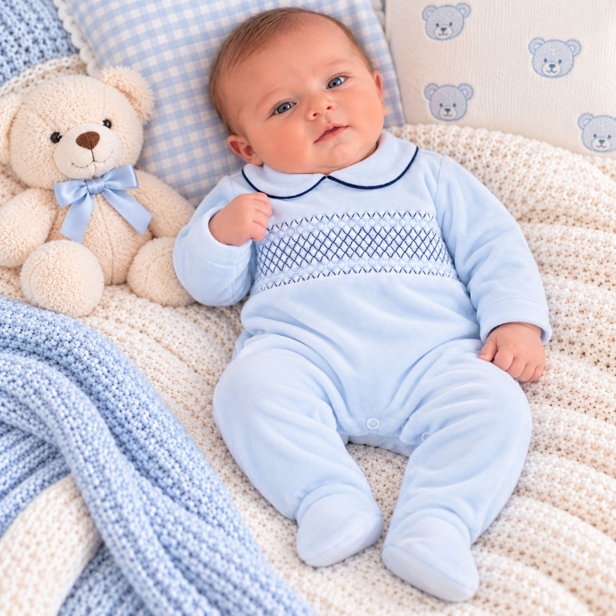 A baby wearing a light blue footed sleeper with a navy blue Peter Pan collar and decorative chest pattern, lying on a cream knit blanket next to a teddy bear and patterned pillows.