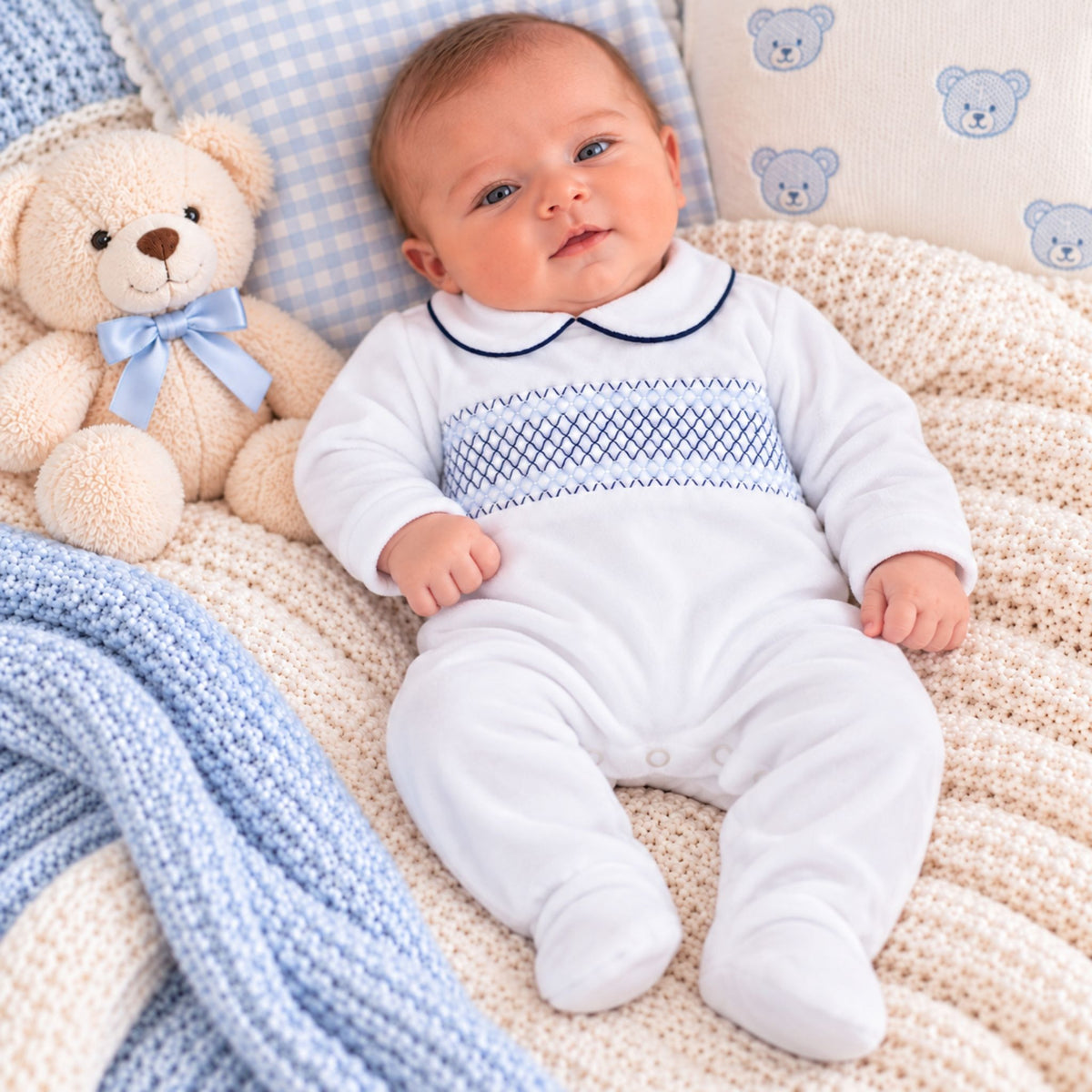 A baby lying on a cream knit blanket next to a blue knit throw, wearing a white footed sleeper with a blue embroidered yoke. A beige teddy bear with a blue bow is on the left, and patterned pillows are in the background.