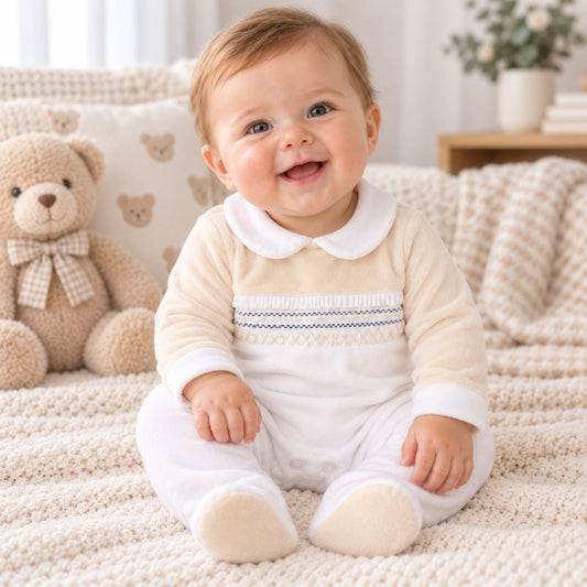 A smiling baby sitting on a textured cream blanket next to a teddy bear, wearing a cream and white long-sleeve footed romper with a Peter Pan collar and a decorative chest stripe.