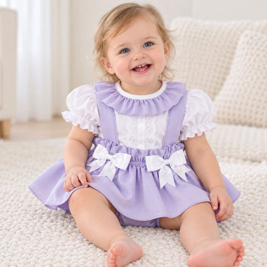 A baby girl sitting on a white textured rug wearing a lavender and white dress. The dress features a lavender pinafore-style bodice with white ruffled sleeves, a white eyelet yoke, and two white bows at the waist.