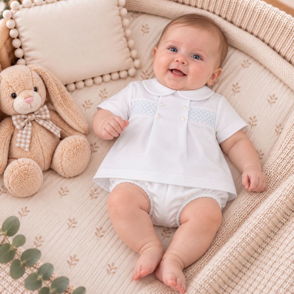 A baby wearing a white short-sleeve collared outfit with a light blue patterned chest panel and white diaper lies on a beige textured blanket in a basket, next to a plush bunny toy.
