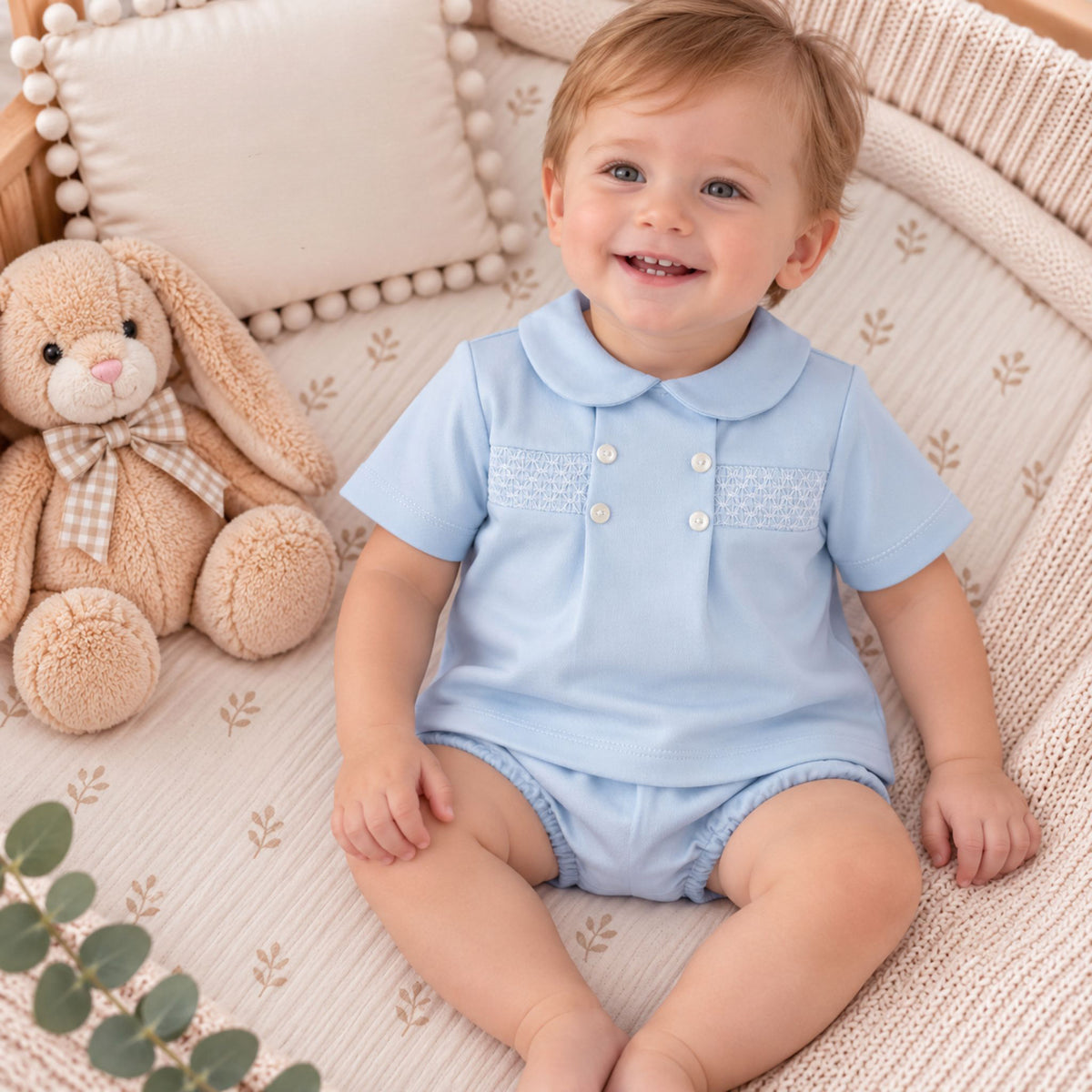 A smiling baby boy wearing a light blue short-sleeve polo romper sits on a cream woven bassinet cushion next to a plush teddy bear.
