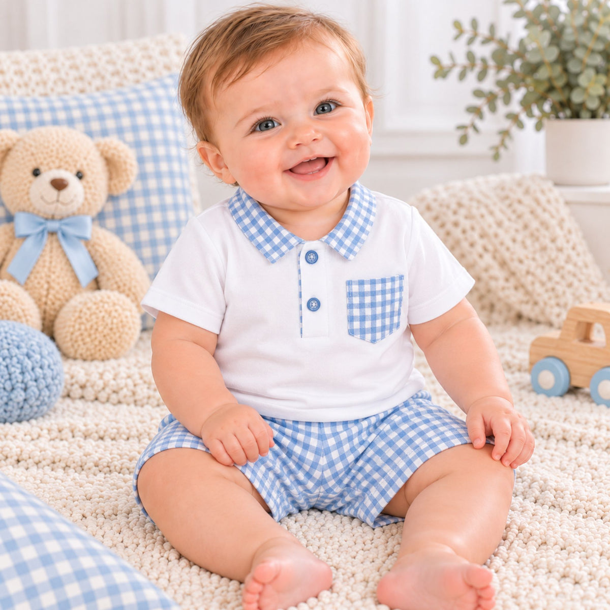 A smiling baby sitting on a cream knit blanket wearing a white short-sleeve polo shirt and matching gingham shorts. The outfit features a blue and white checkered collar and chest pocket.