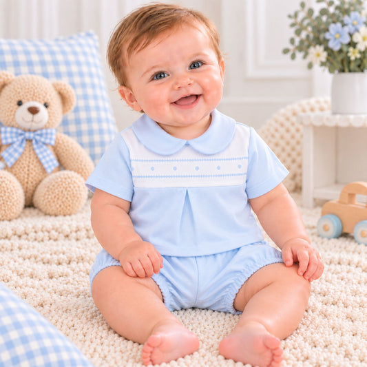 A smiling baby sitting on a cream carpet wearing a light blue short-sleeve romper with a white collar and white chest stripe. A beige teddy bear with a blue gingham bow is on a blue gingham pillow to the left, and a small wooden toy car is on a white side table to the right.