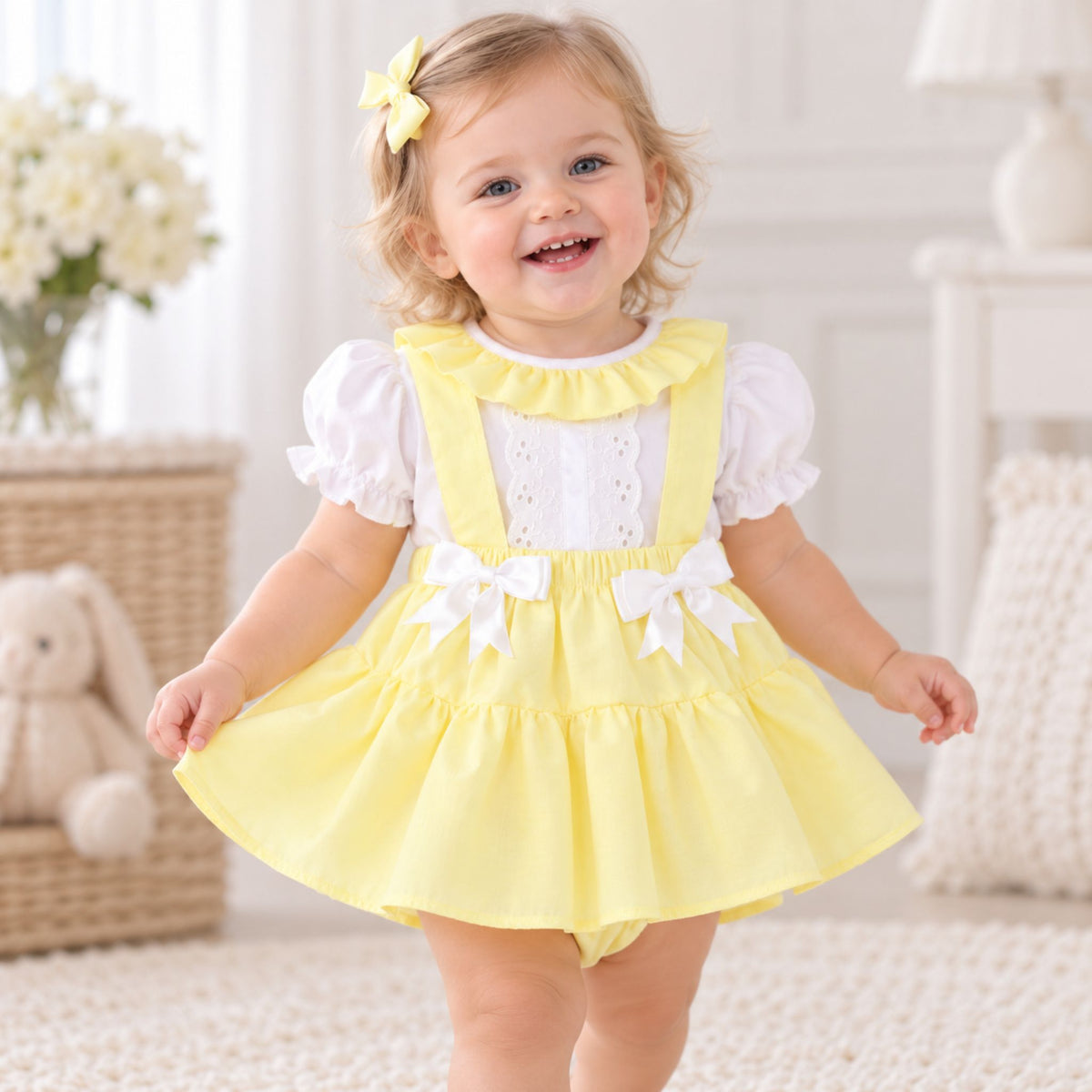 A toddler girl wearing a yellow and white baby dress, smiling in a bright, soft-lit nursery setting.