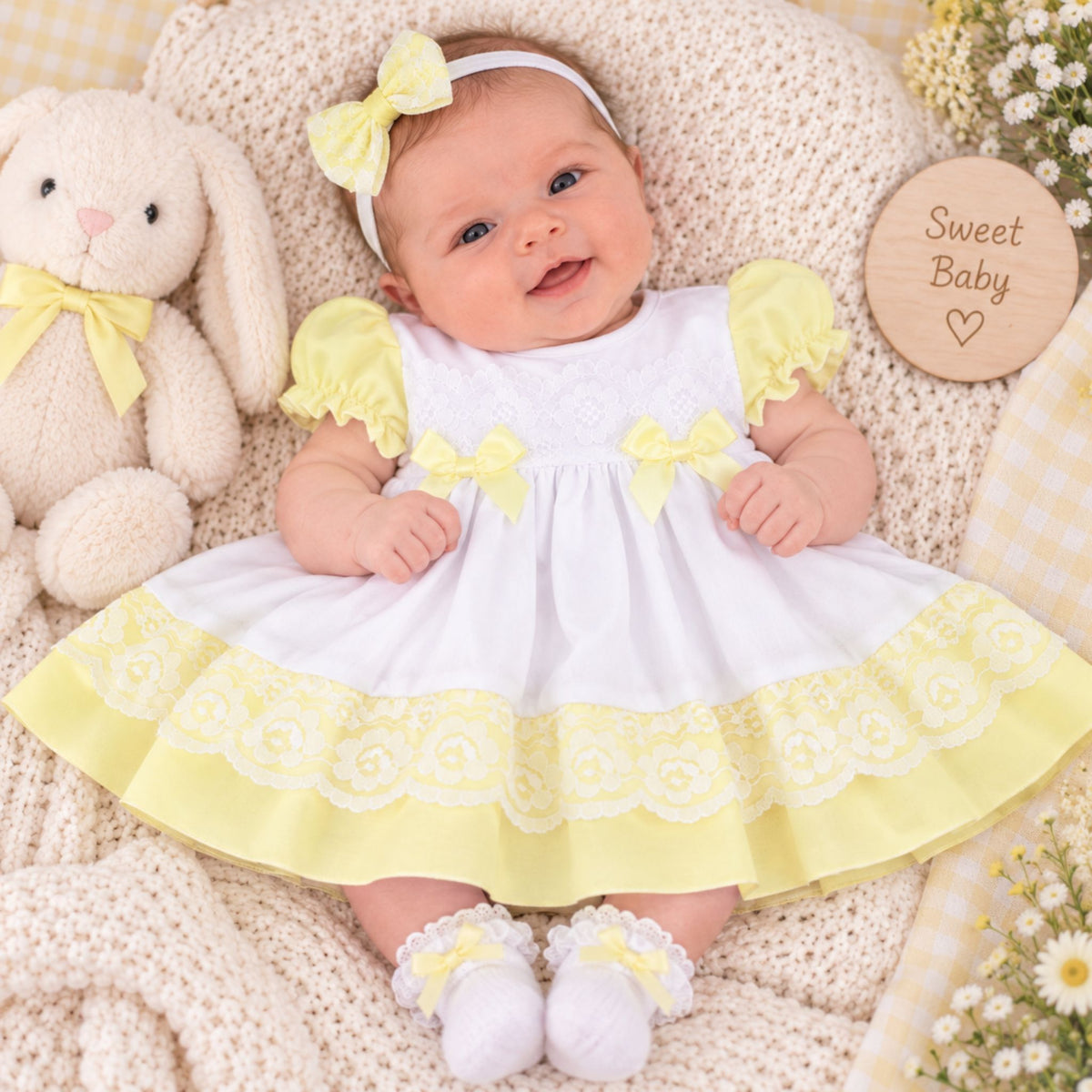 A baby wearing a white and yellow dress with matching yellow bow headband and white booties, lying on a cream knit blanket next to a plush bunny and a round wooden sign that reads 'Sweet Baby'.