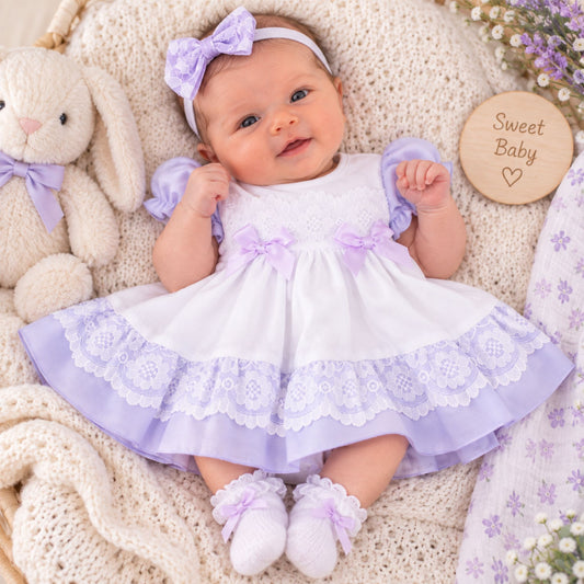 A baby lying on a cream knit blanket wearing a white and lavender baby dress with matching headband and booties, next to a plush bunny and a wooden sign that reads 'Sweet Baby'.