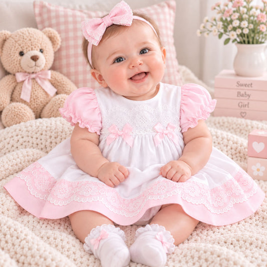 A smiling baby girl wearing a white and pink baby dress with matching pink booties and a pink bow headband, sitting on a cozy cream knit blanket with a teddy bear, pillows, and flowers in the background.