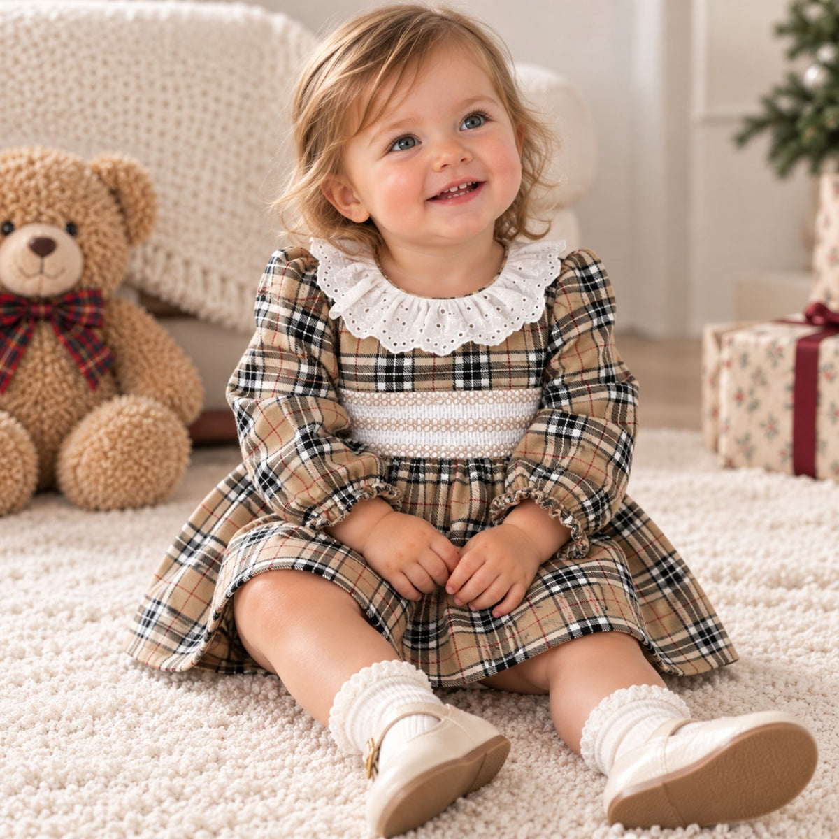 A toddler girl sitting on a cream carpet wearing a tan and black plaid dress with a white lace collar and white knit socks. A teddy bear is visible in the background on the left.