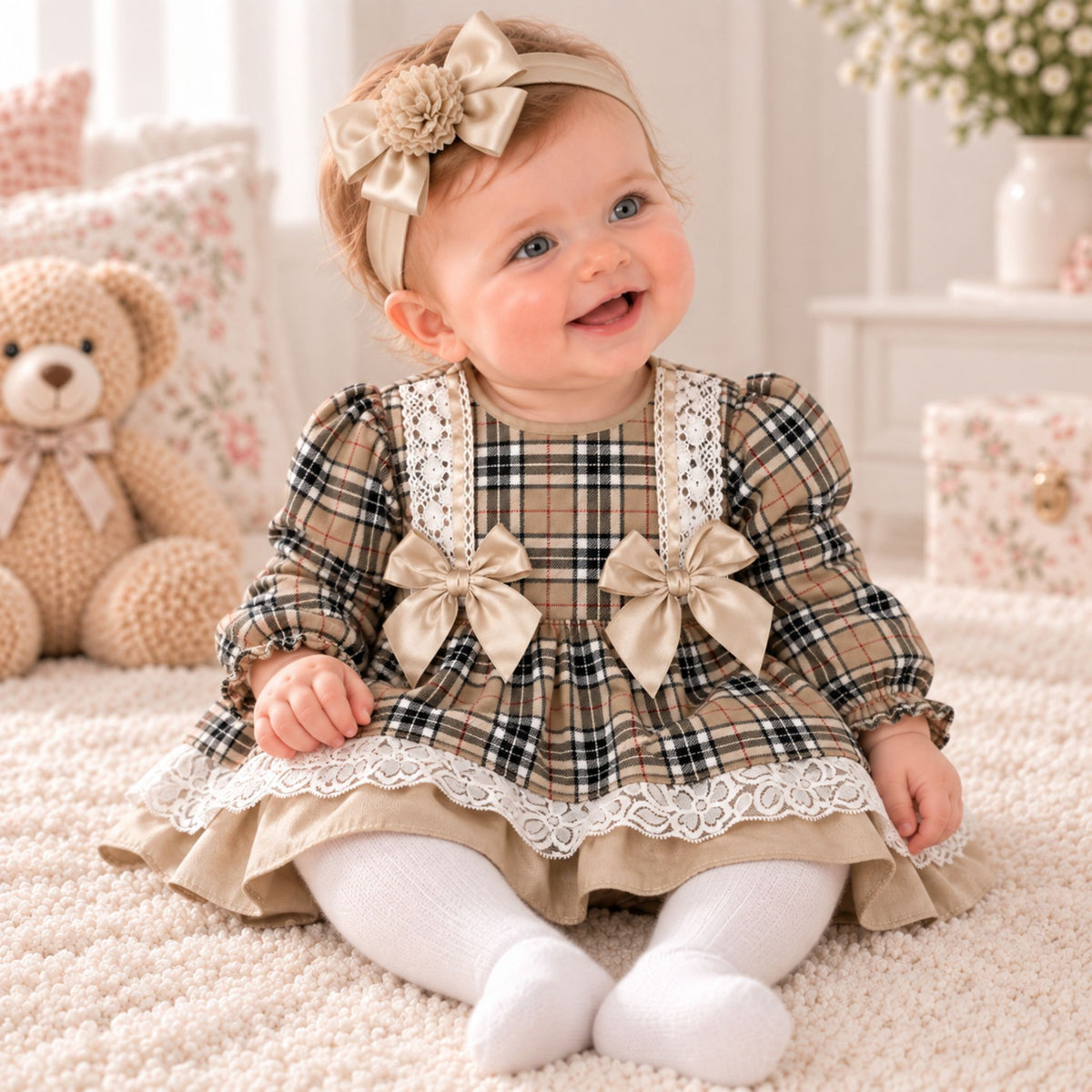 A baby girl sitting on a cream carpet wearing a beige and black plaid dress with white lace trim, matching beige bow details, white tights, and a beige floral headband. A plush teddy bear and soft home decor are visible in the background.