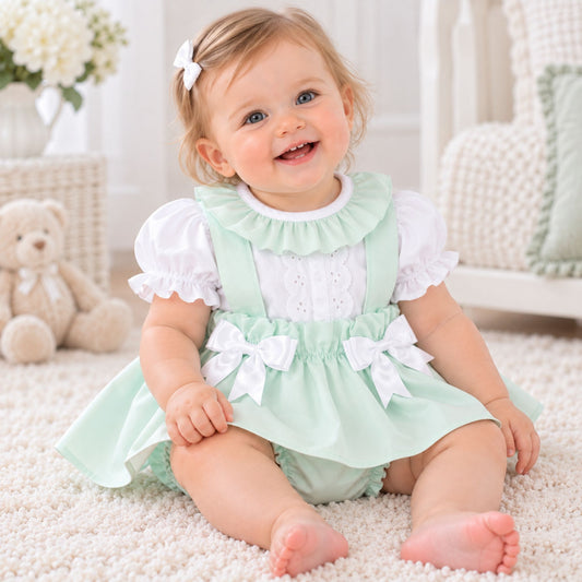 A baby girl sitting on a white carpet wearing a mint green and white dress with ruffles and bow details, in a bright nursery setting.