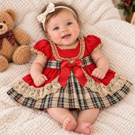 A baby girl wearing a festive red and plaid dress with lace trim and a matching headband, lying on a textured cream blanket next to a teddy bear.