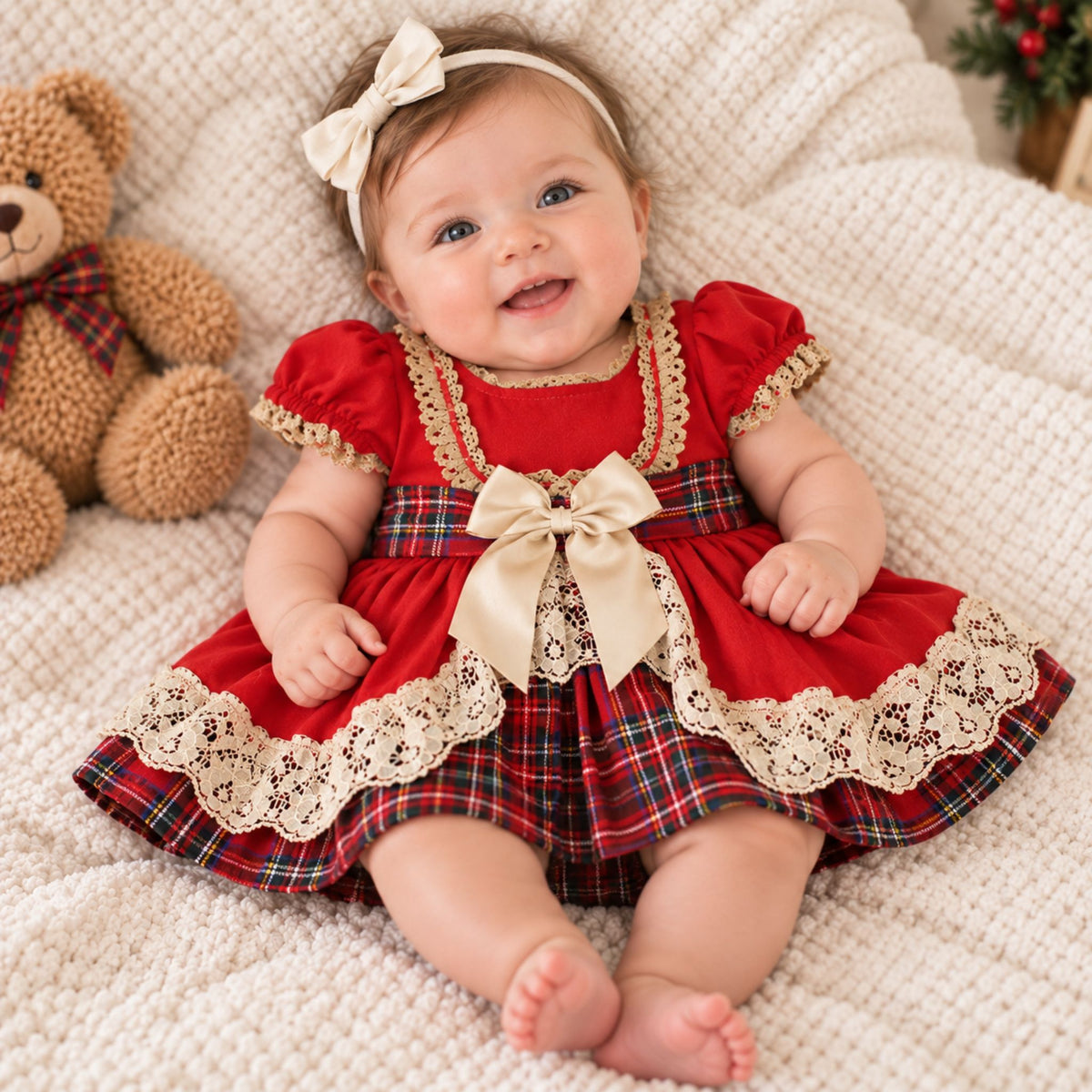 A baby wearing a red holiday-themed dress with a plaid pattern, cream lace trim, and a large cream bow, lying on a textured cream blanket next to a teddy bear.