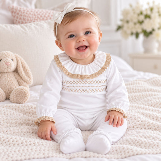 A smiling baby girl wearing a white smocked footed romper with beige lace trim and a matching white bow headband, sitting on a cozy cream knit blanket with a plush bunny toy beside her.