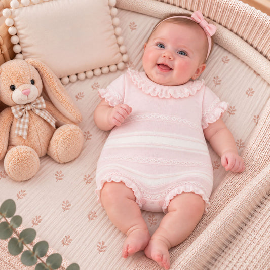 A baby wearing a light pink short-sleeve romper with ruffle trim and a matching pink bow headband, lying in a woven bassinet next to a stuffed bunny toy.
