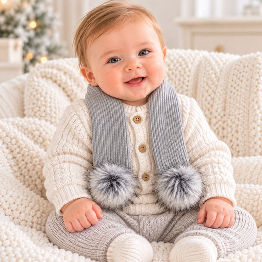 A baby wearing a cream knit sweater, light gray knit pants, and a gray knit scarf with gray faux-fur pom-poms, sitting on a chunky cream knit blanket.