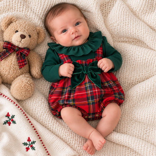 A baby lying on a cream knit blanket next to a teddy bear, wearing a dark green long-sleeve top with a ruffled collar and a red tartan plaid romper with a matching green bow.