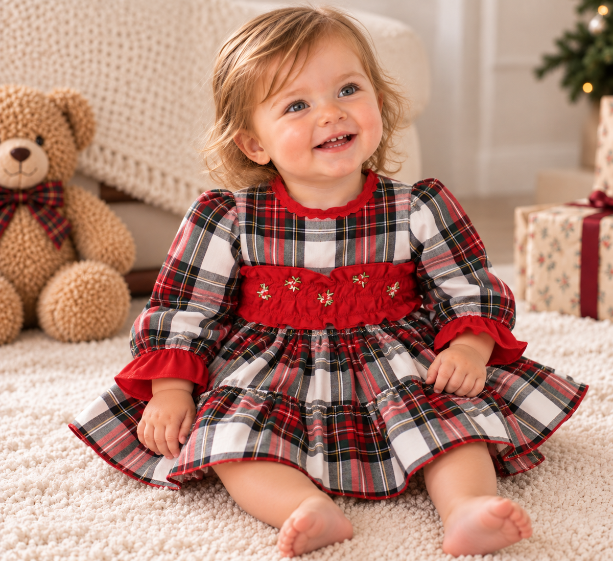 A toddler girl sitting on a cream carpet wearing a red, black, and white plaid dress with a red smocked bodice and gold embroidered details. A brown teddy bear is sitting to her left, and wrapped gift boxes are visible in the background.