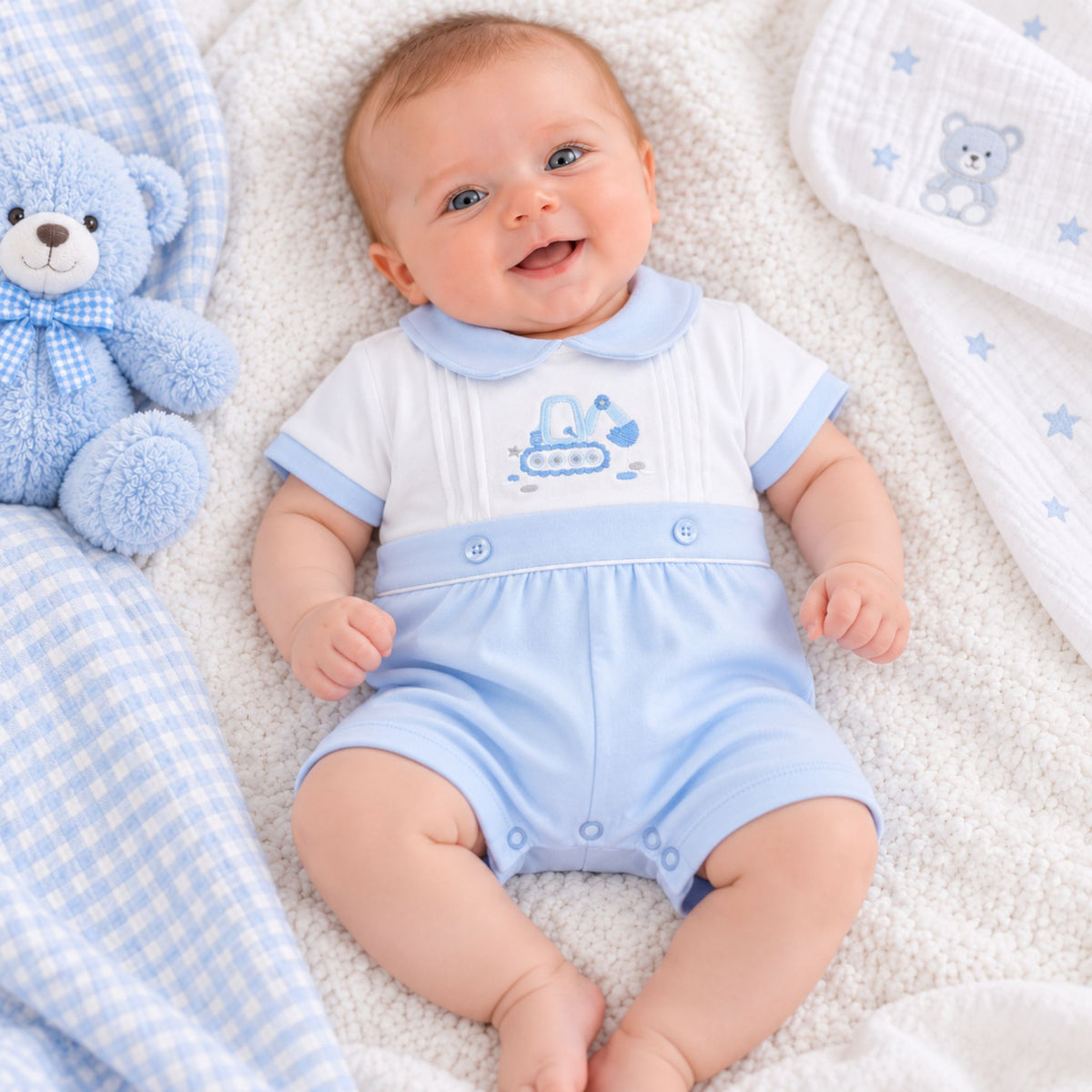 A smiling baby lying on a white textured blanket, wearing a light blue and white short-sleeve romper with a blue boat graphic. A blue teddy bear toy and matching blue-and-white blankets are placed around the baby.