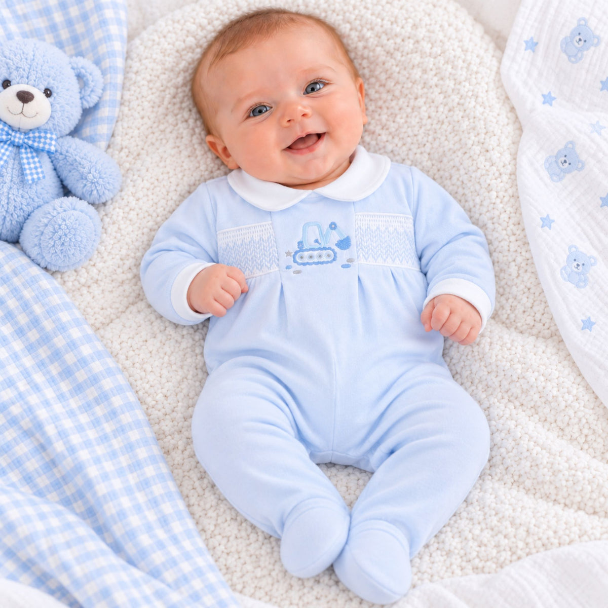A smiling baby lying on a textured cream blanket, wearing a light blue footed onesie with a white collar and a blue submarine graphic on the chest. A blue teddy bear and blue-and-white checkered fabric are visible around the baby.