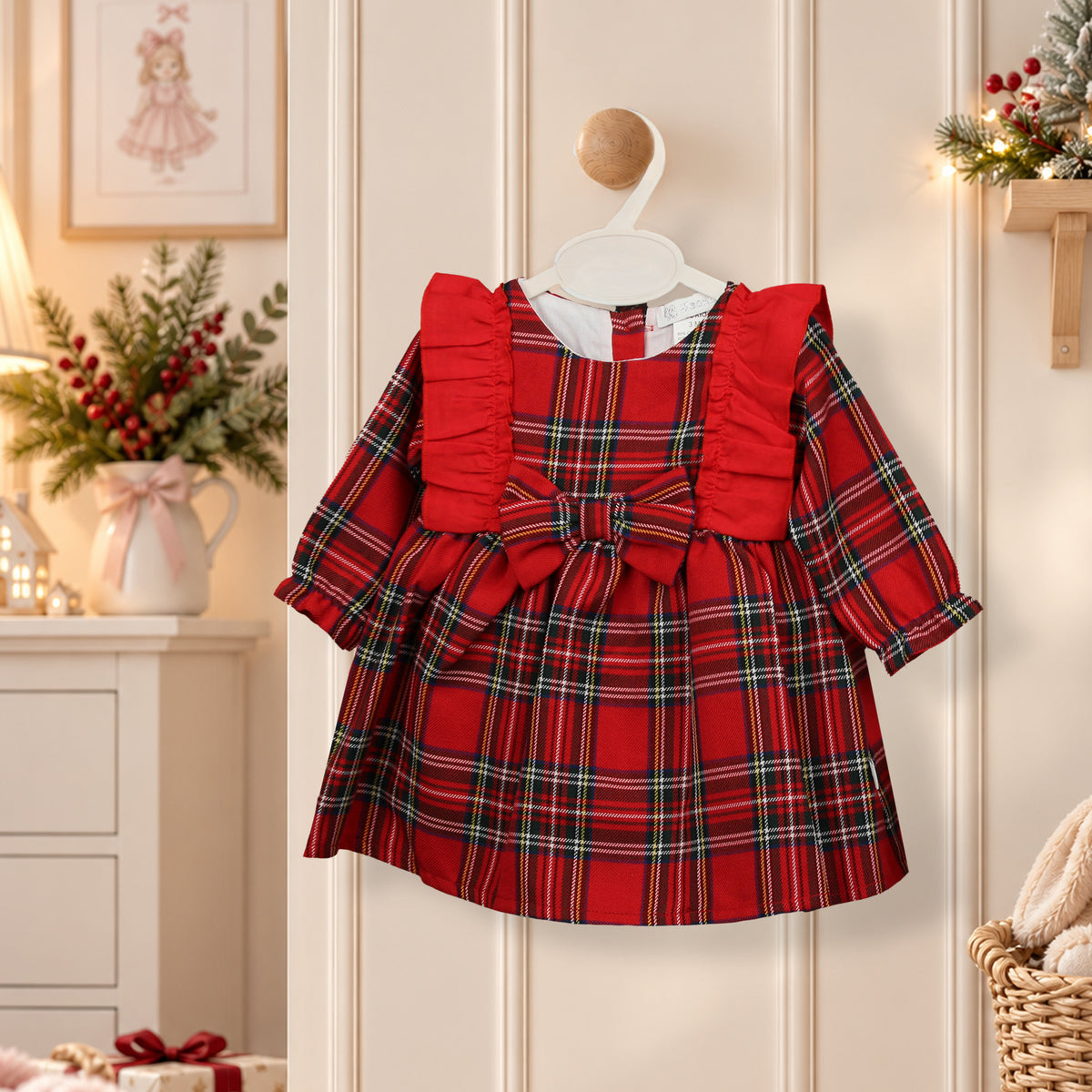 A toddler girl's red plaid dress with ruffled shoulders and a large front bow, hanging on a white hanger against a cream paneled door. The dress features a red, green, and black tartan pattern.