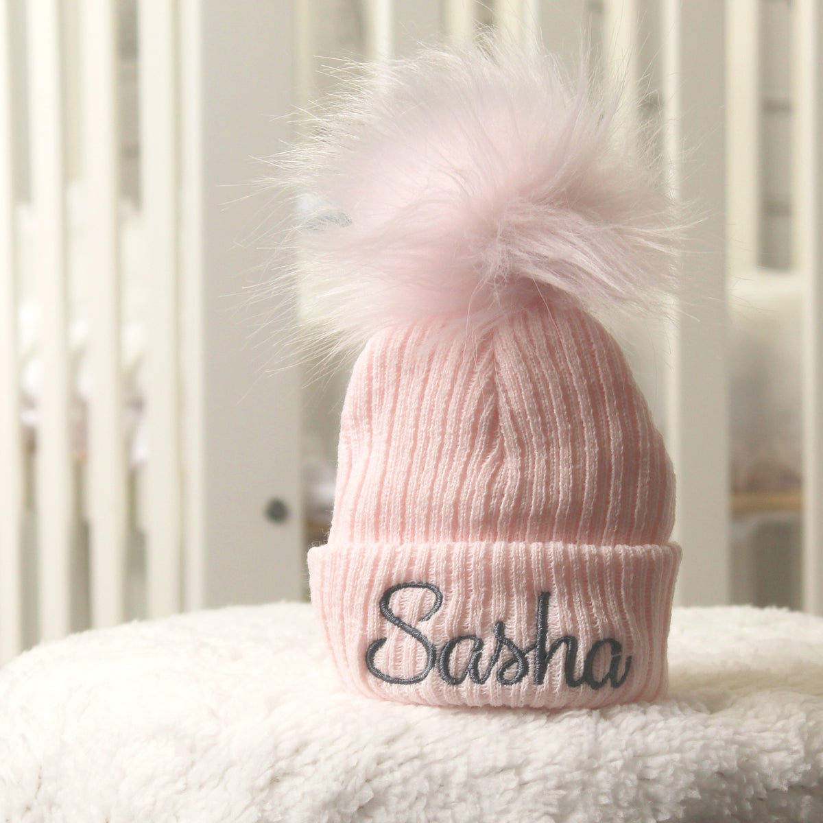 A light pink ribbed knit beanie with a fluffy matching pom-pom on top and the name 'Sasha' embroidered in black script on the front cuff, displayed on a white textured surface.