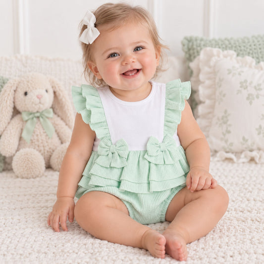 A smiling baby girl sitting on a white textured blanket, wearing a mint green ruffled romper over a white t-shirt. She has a white bow in her hair, with a plush bunny toy and decorative pillows in the background.