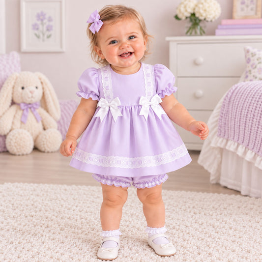 A toddler girl wearing a lavender baby romper dress with white lace trim and bow details, standing in a decorated baby bedroom.