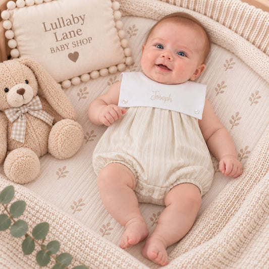 A baby lying on a cream knitted bassinet cushion, wearing a cream striped romper with a white bib personalized with the name 'Joseph'. A plush teddy bear and a decorative pillow reading 'Lullaby Lane Baby Shop' are beside the baby.
