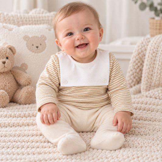 A smiling baby sitting on a cream knit blanket, wearing a beige and white striped long-sleeve top with a white bib and cream footed pants. A teddy bear and soft nursery decor are visible in the background.