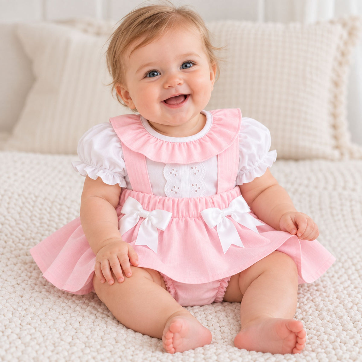 A baby girl sitting on a textured cream knit blanket, wearing a pink and white ruffled baby dress with white bows and white eyelet embroidery on the chest.