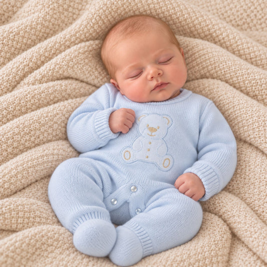 A sleeping newborn baby wearing a light blue knit footed romper with an embroidered teddy bear on the chest, lying on a beige textured blanket.