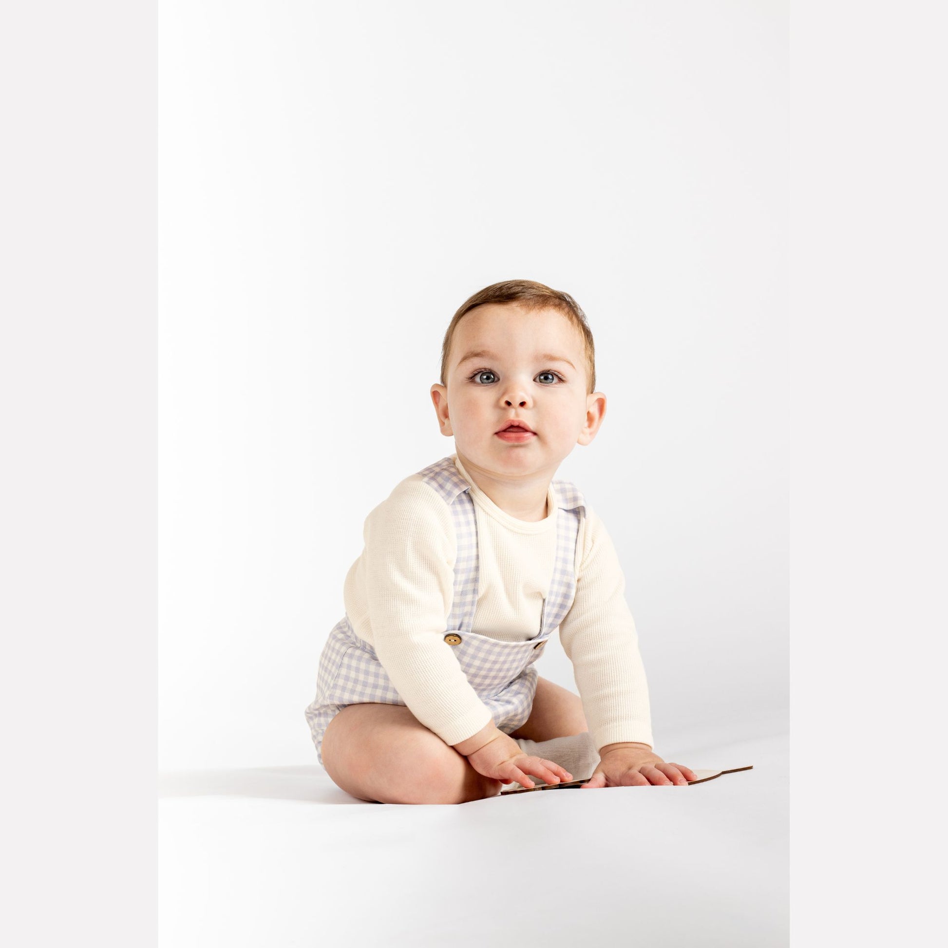 Baby sitting on a white surface wearing a white shirt and checkered overalls.