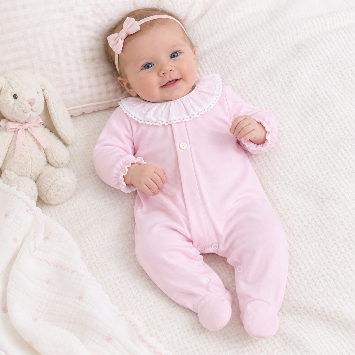 A baby lying on a soft cream blanket wearing a light pink footed sleeper with a ruffled white collar and a matching pink bow headband. A plush bunny toy sits beside the baby.