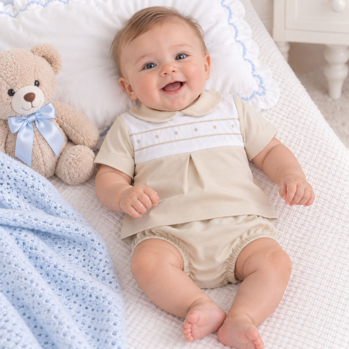 A smiling baby lying on a white textured blanket next to a teddy bear, wearing a beige short-sleeve romper with a white chest panel and embroidered details.