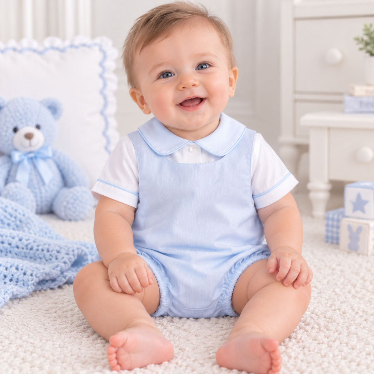 A smiling baby sitting on a white textured rug, wearing a light blue overall romper over a white short-sleeve shirt. The background features a soft blue teddy bear, a blue crocheted blanket, and a white dresser.