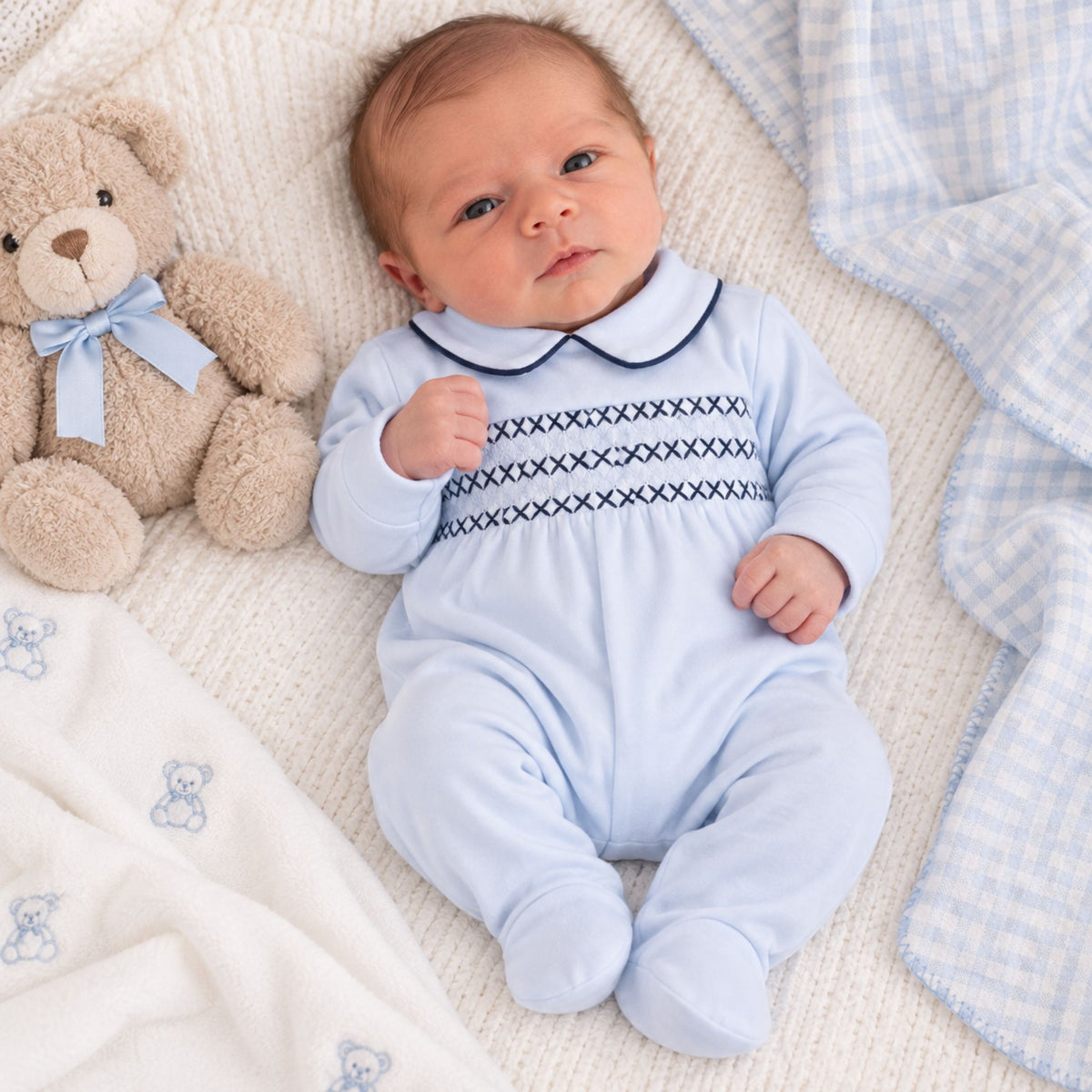 A baby lying on a cream knit blanket next to a teddy bear, wearing a light blue footed onesie with dark blue embroidered horizontal lines across the chest.